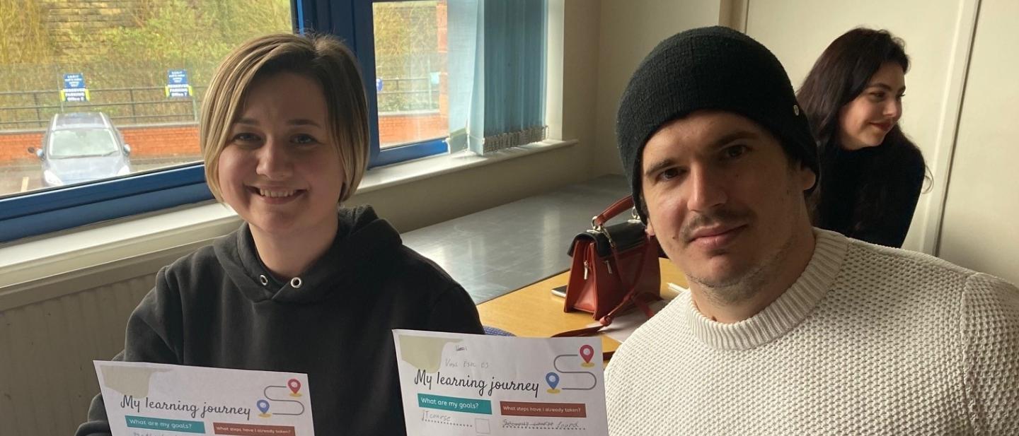 Two people are sat at a desk and are holding up their learning plans
