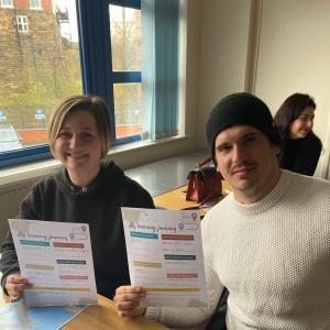Two people are sat at a desk and are holding up their learning plans