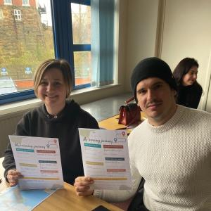 Two people are sat at a desk and are holding up their learning plans